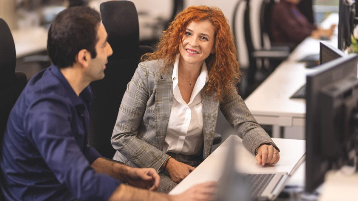 a woman and a man in an office sitting at a desk with a computer on it